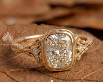 May include: A gold ring featuring a central cushion-cut gemstone surrounded by a beaded bezel setting. The band displays intricate scrollwork engravings and is accented with two small round stones on either side of the centre setting.