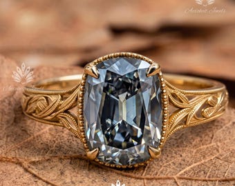 May include: A gold ring featuring a large, cushion-cut, grey gemstone set in a beaded bezel. The band is crafted from yellow gold and displays intricate, raised leaf-like engravings along the shoulders, extending from the central setting.