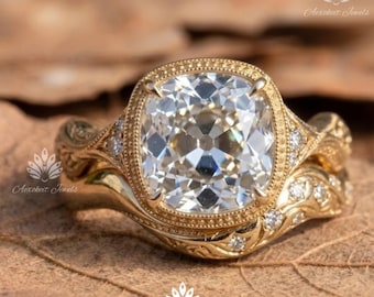 May include: A gold ring featuring a large, cushion-cut center stone surrounded by a milgrain-detailed bezel. The band is crafted with intricate, swirling filigree patterns and is accented with small, round, clear stones along the shoulders.