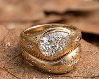 Puede incluir: Un anillo de oro con una piedra central en forma de pera y múltiples grabados en forma de estrella, cada uno acentuado con un pequeño diamante incrustado. La banda tiene un acabado liso y pulido con un perfil ancho y redondeado.