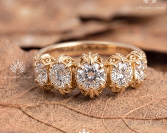 May include: A gold ring featuring five round, clear, faceted stones set in a row. The metal band displays intricate, decorative metalwork surrounding each stone, creating a continuous, detailed pattern across the front of the ring.