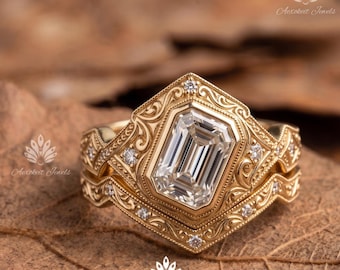 Puede incluir: Un conjunto de anillos de oro con una piedra central de corte esmeralda en engaste de bisel, rodeada de intrincados grabados de volutas y pequeños diamantes redondos. La banda tiene una forma contorneada a juego con detalles metálicos.