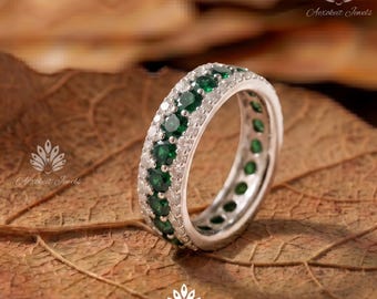 May include: A silver-toned eternity band featuring a central row of round-cut dark green gemstones flanked by two rows of small, clear, sparkling stones. The ring has a polished metal finish and a continuous design of stones around the entire band.