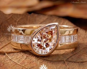 May include: A gold ring featuring a large pear-shaped champagne-coloured centre stone set in a bezel, flanked by a row of channel-set baguette-cut clear stones along the band. The design includes a polished gold finish and a wide band structure.