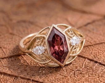 Puede incluir: Un anillo de oro con una gema central de color marrón rojizo en forma de marquesa, flanqueada por dos gemas blancas más pequeñas. El anillo tiene un diseño intrincado en forma de hoja. El anillo está sobre una superficie marrón texturizada.