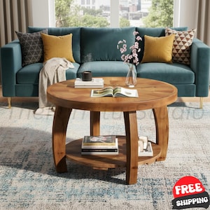 May include: A round wooden coffee table with a natural grain finish, featuring a circular lower shelf for storage. The table has four curved legs and supports an open book on its surface, along with a glass vase containing pink floral stems.