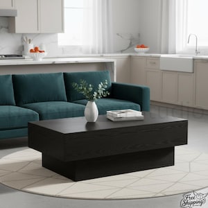 May include: A rectangular black wooden coffee table with a thick, tiered base design. The table features a visible wood grain texture and is positioned on a round, light-colored area rug in front of a deep teal sofa with a white vase and books.