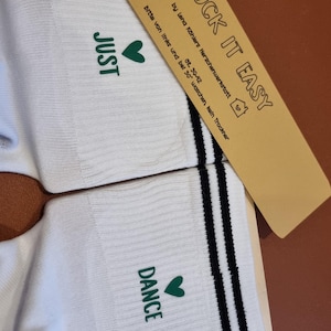 Pu&ograve; includere: Calzini bianchi con le parole "JUST" e "DANCE" stampate in verde, ciascuna accompagnata da un cuore verde. I calzini hanno strisce nere e il testo "SOCK IT EASY" su un'etichetta marrone.