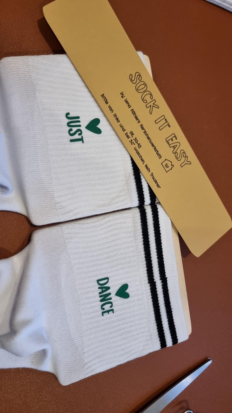 Pu&ograve; includere: Calzini bianchi con le parole "JUST" e "DANCE" stampate in verde, ciascuna accompagnata da un cuore verde. I calzini hanno strisce nere e il testo "SOCK IT EASY" su un'etichetta marrone.