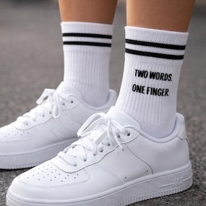 Può includere: Scarpe sportive bianche con calzini bianchi. I calzini hanno due strisce nere in alto e il testo "TWO WORDS. ONE FINGER." Le scarpe sono allacciate e hanno una suola strutturata.