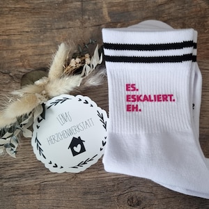 Può includere: Calzini bianchi con strisce nere e la scritta rosa "ES. ESKALIERT. EH.". Un'etichetta con la scritta "LENAS HERZCHENWERKSTATT" e un'icona a forma di casa è accanto ai calzini. Sono presenti anche fiori secchi.