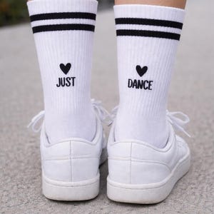 May include: White socks with black stripes and the words "JUST DANCE" in black. The socks are worn with white sneakers. The image is a close-up of the feet and ankles.