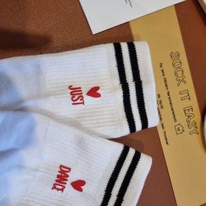 Pu&ograve; includere: Calzini bianchi con strisce nere e le parole "JUST" e "DANCE" in rosso, ciascuna accompagnata da un cuore rosso. I calzini sono su una superficie marrone, con una carta "SOCK IT EASY".