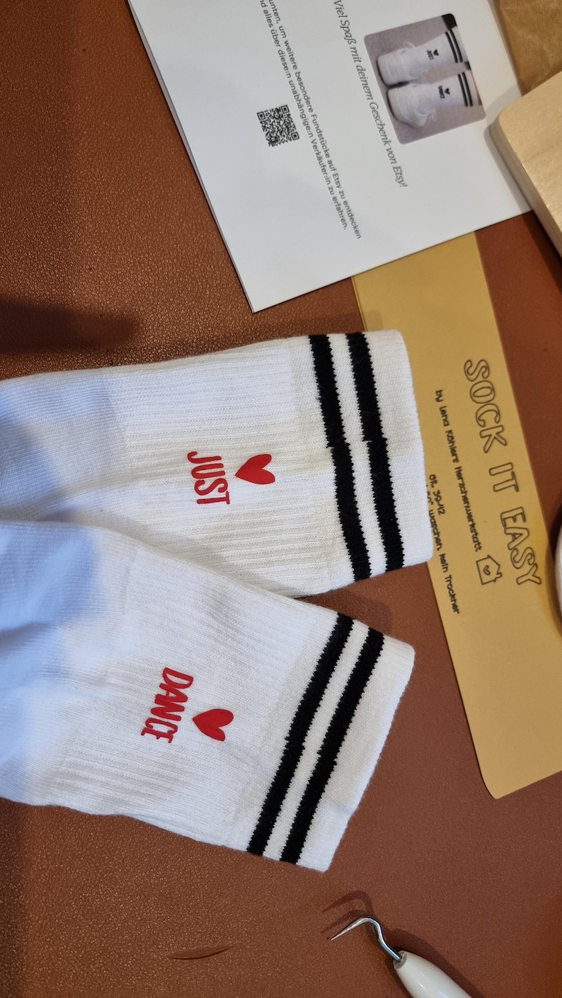 Pu&ograve; includere: Calzini bianchi con strisce nere e le parole "JUST" e "DANCE" in rosso, ciascuna accompagnata da un cuore rosso. I calzini sono su una superficie marrone, con una carta "SOCK IT EASY".