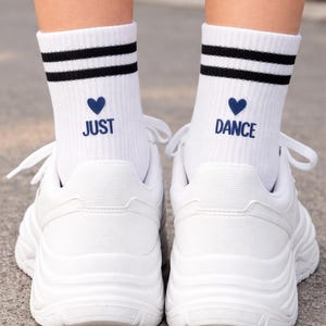 Pu&ograve; includere: Scarpe da ginnastica bianche con suole spesse abbinate a calzini bianchi. I calzini hanno due strisce nere, un cuore blu e le parole "JUST DANCE" in blu.