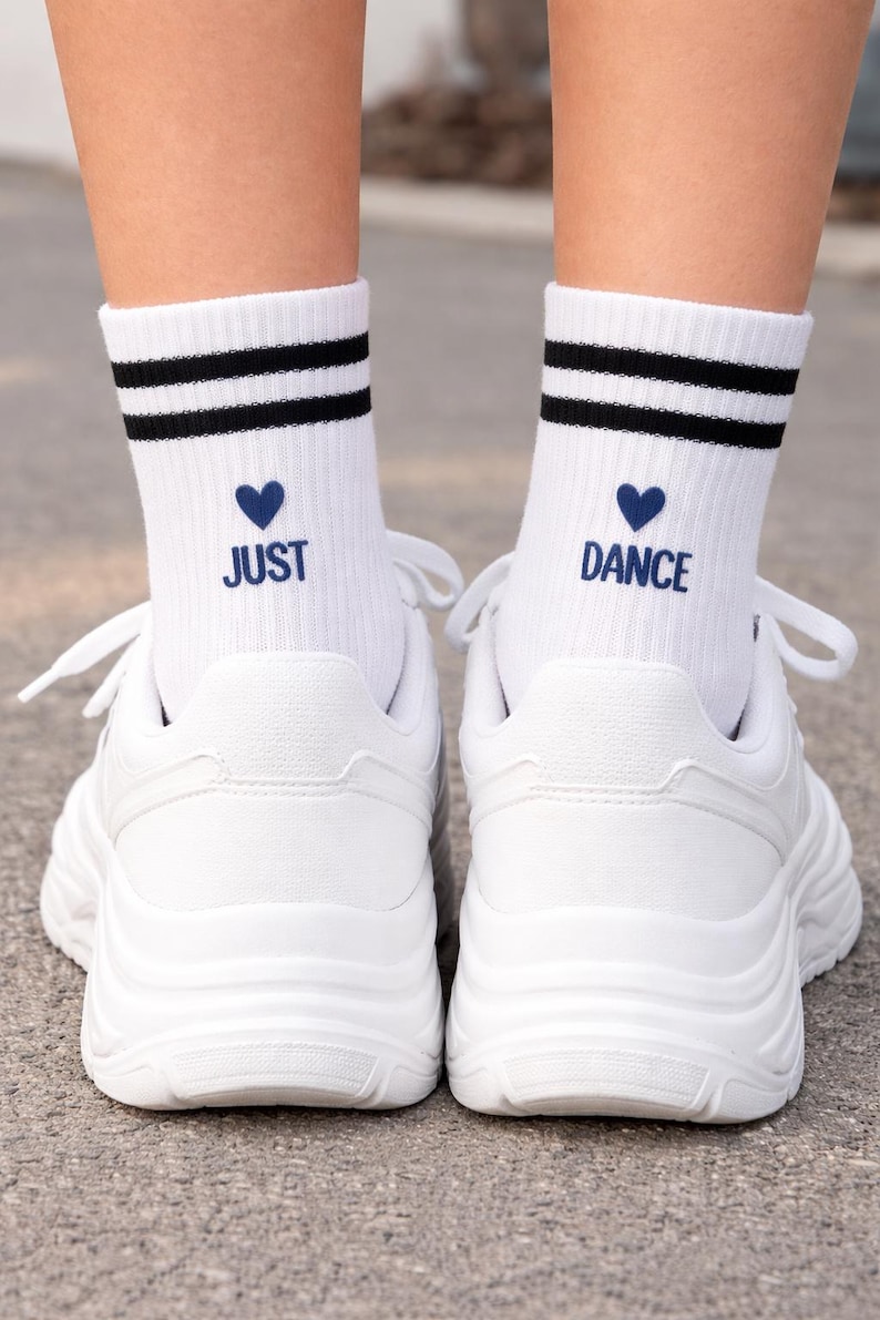 Pu&ograve; includere: Scarpe da ginnastica bianche con suole spesse abbinate a calzini bianchi. I calzini hanno due strisce nere, un cuore blu e le parole "JUST DANCE" in blu.