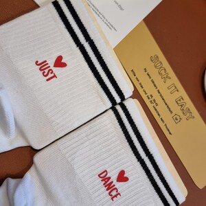 Pu&ograve; includere: Un paio di calzini bianchi con strisce nere e le parole "JUST" e "DANCE" in rosso, ognuna con un cuore rosso. I calzini sono su una superficie marrone con una carta che dice "SOCK IT EASY".