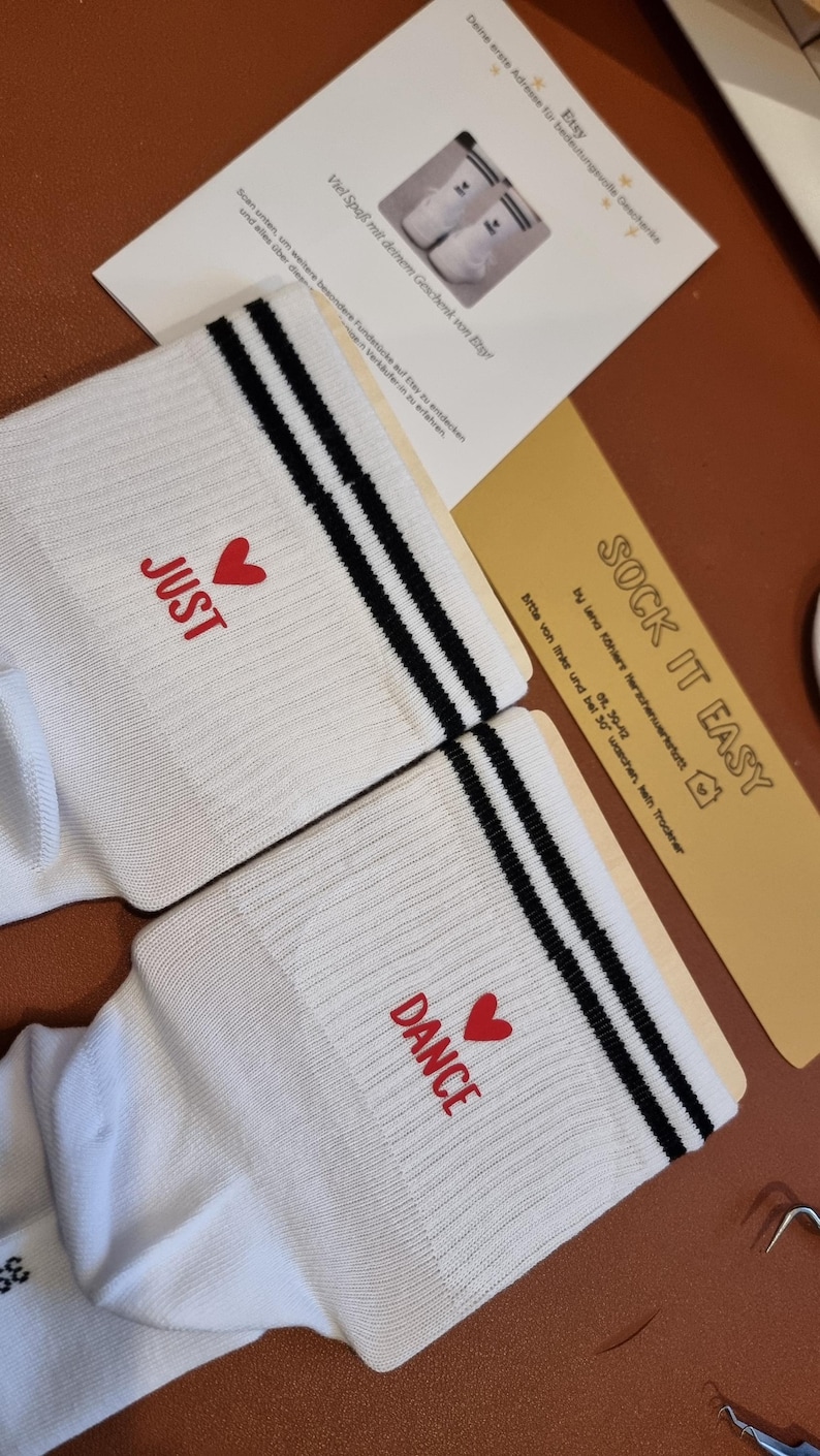 Pu&ograve; includere: Un paio di calzini bianchi con strisce nere e le parole "JUST" e "DANCE" in rosso, ognuna con un cuore rosso. I calzini sono su una superficie marrone con una carta che dice "SOCK IT EASY".