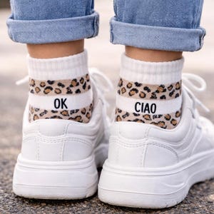 以下が含まれることがあります： ヒョウ柄のバンドが付いた白い靴下で、「OK」と「CIAO」の文字がプリントされています。白いスニーカーと青いジーンズに合わせています。