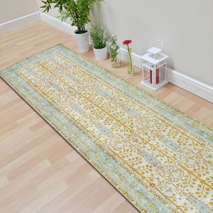 May include: A rectangular runner rug with a cream-colored background and a repeating pattern of stylized trees in gold and blue. The rug has a light blue border and is placed on a light wood floor.