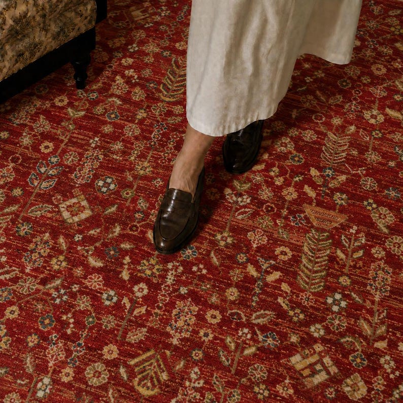Peut inclure: Un tapis oriental rouge avec un motif floral. Le tapis pr&eacute;sente un design d&eacute;taill&eacute; avec diverses fleurs et feuilles dans des tons de jaune, vert et bleu. Une personne porte des mocassins en cuir noir et une robe blanche.