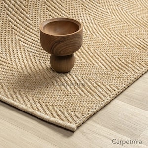 May include: A woven beige rug with a chevron pattern. A small, wooden bowl sits on the rug. The rug is made of natural fibers and has a textured appearance. The rug is on a light-colored wooden floor.