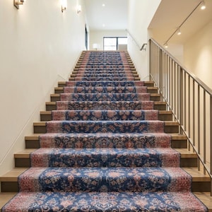 May include: A staircase with wooden steps and a runner rug. The rug features a detailed floral pattern in shades of blue and red. The walls are painted a light beige color, and there is a metal railing on the right side.