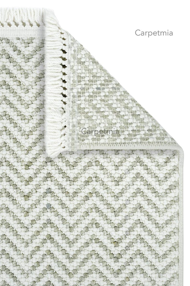 May include: A close-up of a white and gray rug with a chevron pattern. The rug has a textured surface and a fringe along one edge. The brand name "Carpetmia" is visible on the rug.