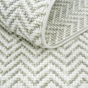 May include: Close-up of a textured rug with a repeating chevron pattern. The rug features a combination of white and light grey colours, with a soft, looped pile. The edge of the rug is finished with a light grey border.