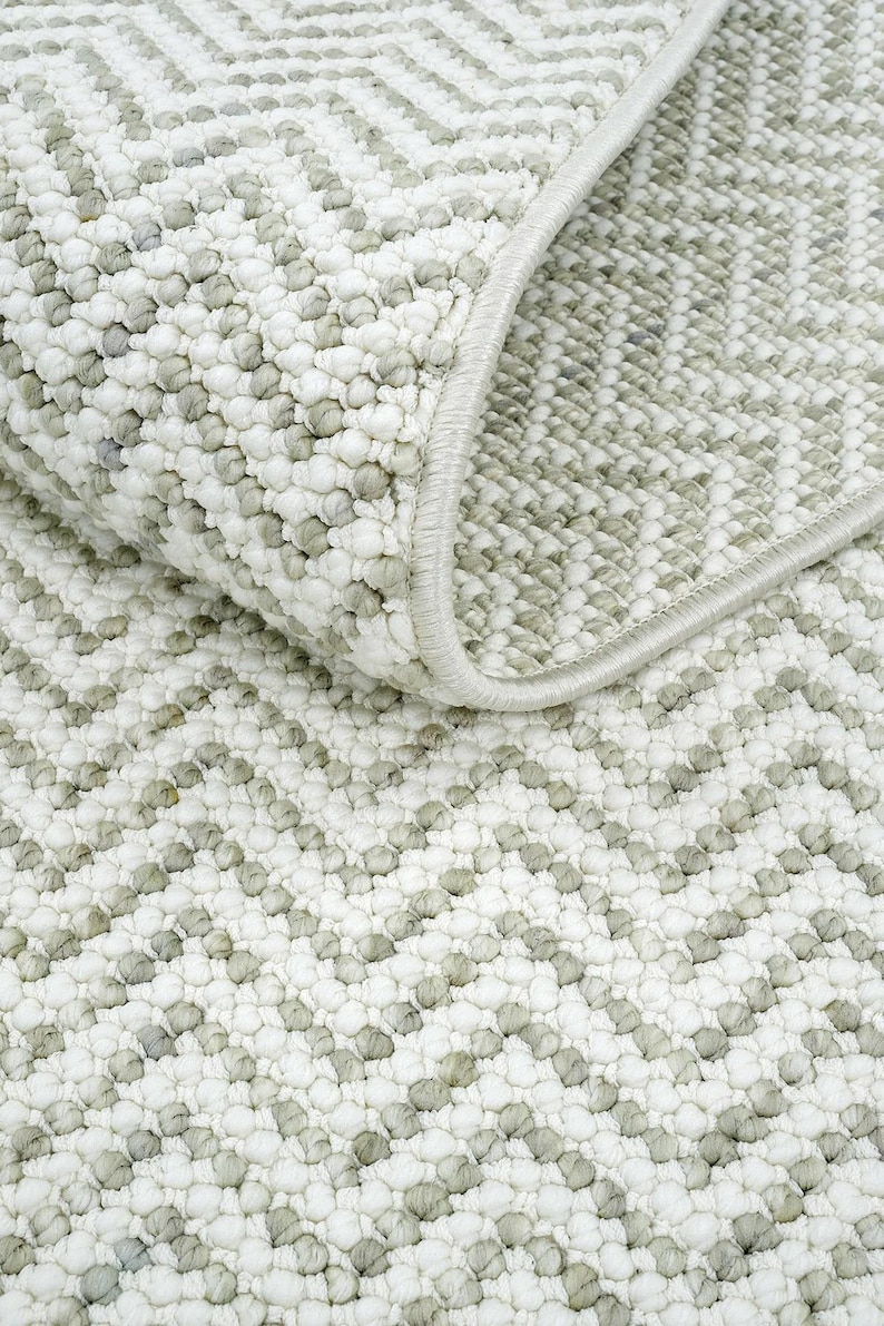 May include: Close-up of a textured rug with a repeating chevron pattern. The rug features a combination of white and light gray colors, with a soft, looped pile. The edge of the rug is finished with a light gray border.
