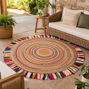 Può includere: Un tappeto rotondo intrecciato con cerchi concentrici di fibre naturali color beige, rosso, giallo, verde e blu. Il bordo esterno è rifinito con una frangia decorativa composta da brevi strisce di tessuto multicolore in tonalità vivaci.