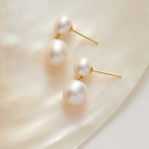 May include: Two pairs of pearl earrings with gold posts rest on a pearlescent, off-white surface. Each earring features two white pearls. The earrings are displayed on a shell-shaped surface, with a soft, diffused light.
