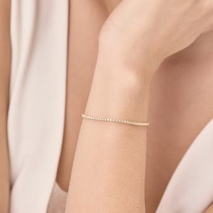 Könnte beinhalten: Ein zartes goldenes Armband, besetzt mit zahlreichen kleinen, funkelnden Edelsteinen. Das Armband wird am Handgelenk getragen und zeigt sein elegantes Design und seinen subtilen Schimmer. Das Schmuckstück ist ein klassisches Accessoire.