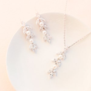 May include: A delicate jewelry set featuring a necklace and earrings. The set includes a silver-toned chain necklace and matching earrings, each adorned with leaf-shaped crystals and pearls. The jewelry is displayed on a white plate.