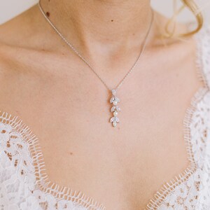 May include: A silver necklace with a pendant featuring a leaf design and small pearls. The necklace is worn against a white lace garment, creating a delicate and elegant aesthetic. The pendant is approximately 2 inches long.