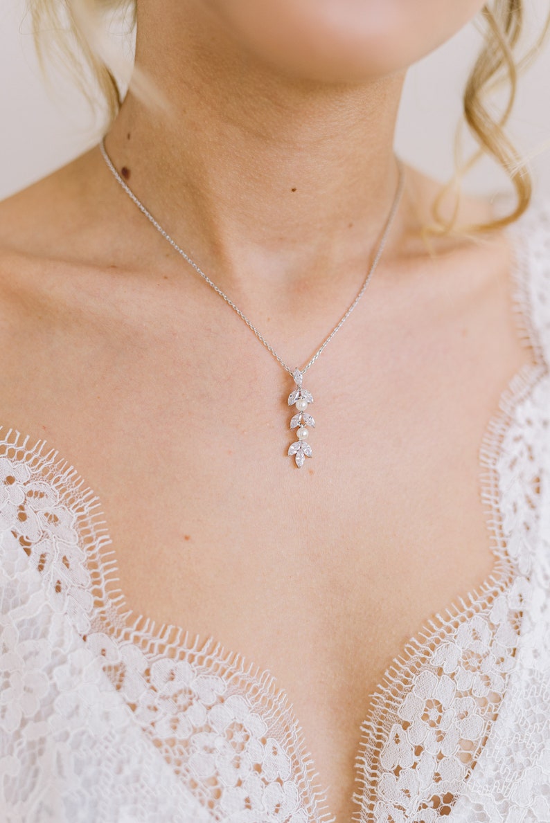 May include: A silver necklace with a pendant featuring a leaf design and small pearls. The necklace is worn against a white lace garment, creating a delicate and elegant aesthetic. The pendant is approximately 2 inches long.