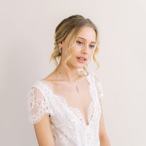 May include: A white lace wedding dress with a plunging neckline and short sleeves. The dress features intricate lace detailing and a jeweled belt. The model is also wearing a matching necklace and earrings. The overall look is elegant and romantic.