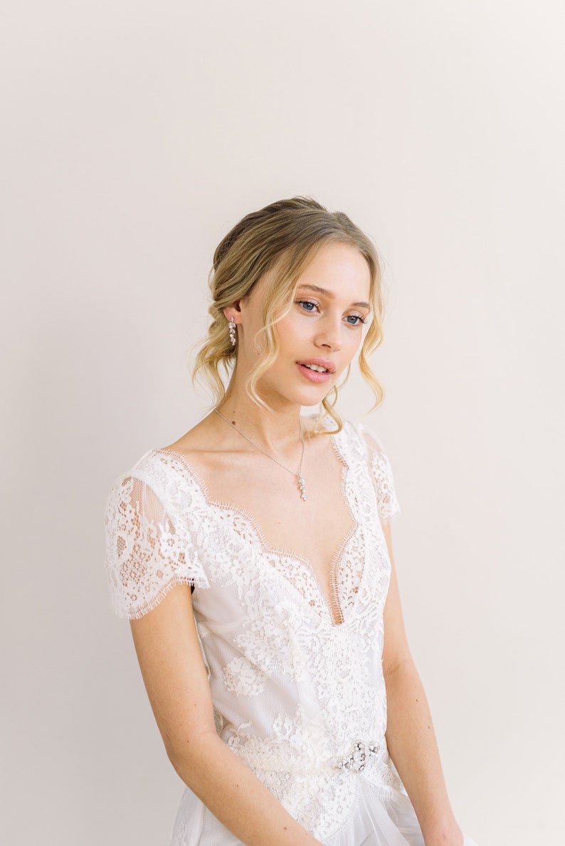 May include: A white lace wedding dress with a plunging neckline and short sleeves. The dress features intricate lace detailing and a jeweled belt. The model is also wearing a matching necklace and earrings. The overall look is elegant and romantic.