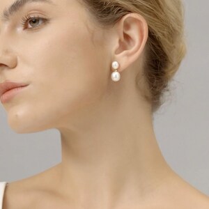 May include: A pair of elegant, gold-tone earrings featuring two white, round pearls. The earrings are displayed on a person's ear, showcasing their classic design and subtle shimmer. The pearls are of different sizes, with a smaller pearl on top.