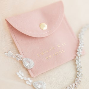 May include: A close-up of bridal jewelry, including a sparkling necklace and teardrop earrings. The earrings and necklace are adorned with clear crystals. A pink pouch with the text "EMILIA RAI BRIDAL" is also in the image.
