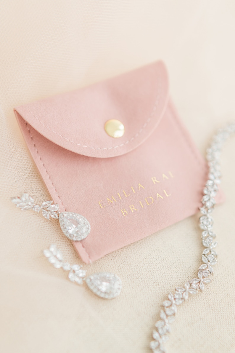 May include: A close-up of bridal jewelry, including a sparkling necklace and teardrop earrings. The earrings and necklace are adorned with clear crystals. A pink pouch with the text "EMILIA RAI BRIDAL" is also in the image.