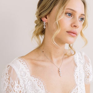May include: A silver necklace and matching earrings set. The jewelry features a delicate leaf design with clear, sparkling stones. The earrings and necklace are worn by a person wearing a white lace dress.