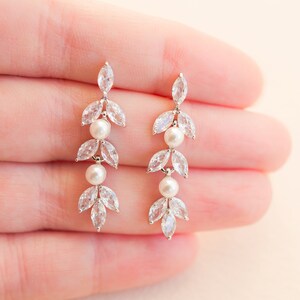 May include: Elegant dangle earrings featuring a leaf design with clear, marquise-cut crystals and two small, round pearls. The earrings have a silver-tone finish and are designed for pierced ears. A delicate and sophisticated accessory.