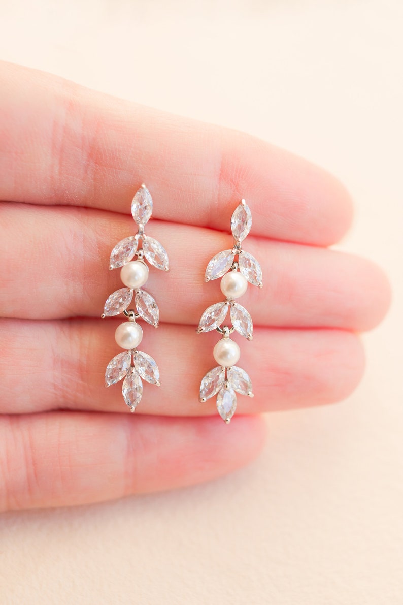 May include: Elegant dangle earrings featuring a leaf design with clear, marquise-cut crystals and two small, round pearls. The earrings have a silver-tone finish and are designed for pierced ears. A delicate and sophisticated accessory.