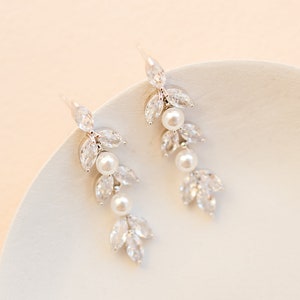 May include: Elegant silver-toned earrings with a leaf and pearl design. Each earring features three pear-shaped clear crystals and three white pearls. The earrings are displayed on a white surface against a soft peach background.