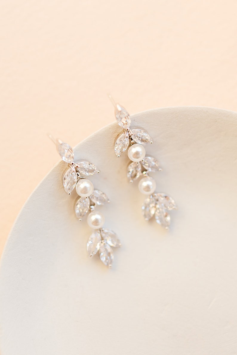 May include: Elegant silver-toned earrings with a leaf and pearl design. Each earring features three pear-shaped clear crystals and three white pearls. The earrings are displayed on a white surface against a soft peach background.
