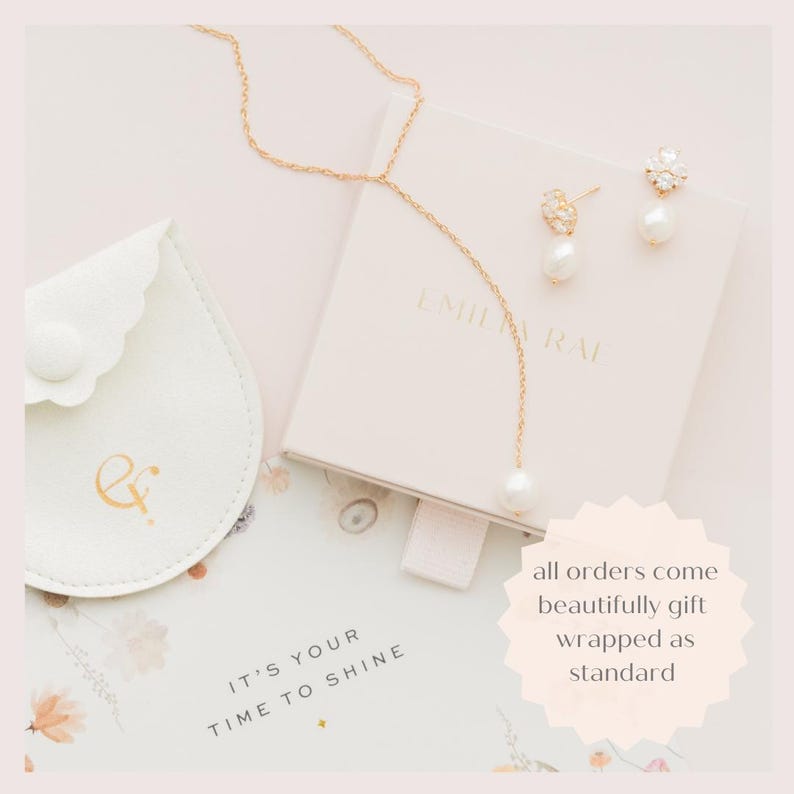 May include: A gold-colored necklace with a single pearl pendant and matching earrings with pearl drops are displayed on a light pink box. A small, white, scalloped pouch with gold lettering and the text "IT'S YOUR TIME TO SHINE" are also visible.