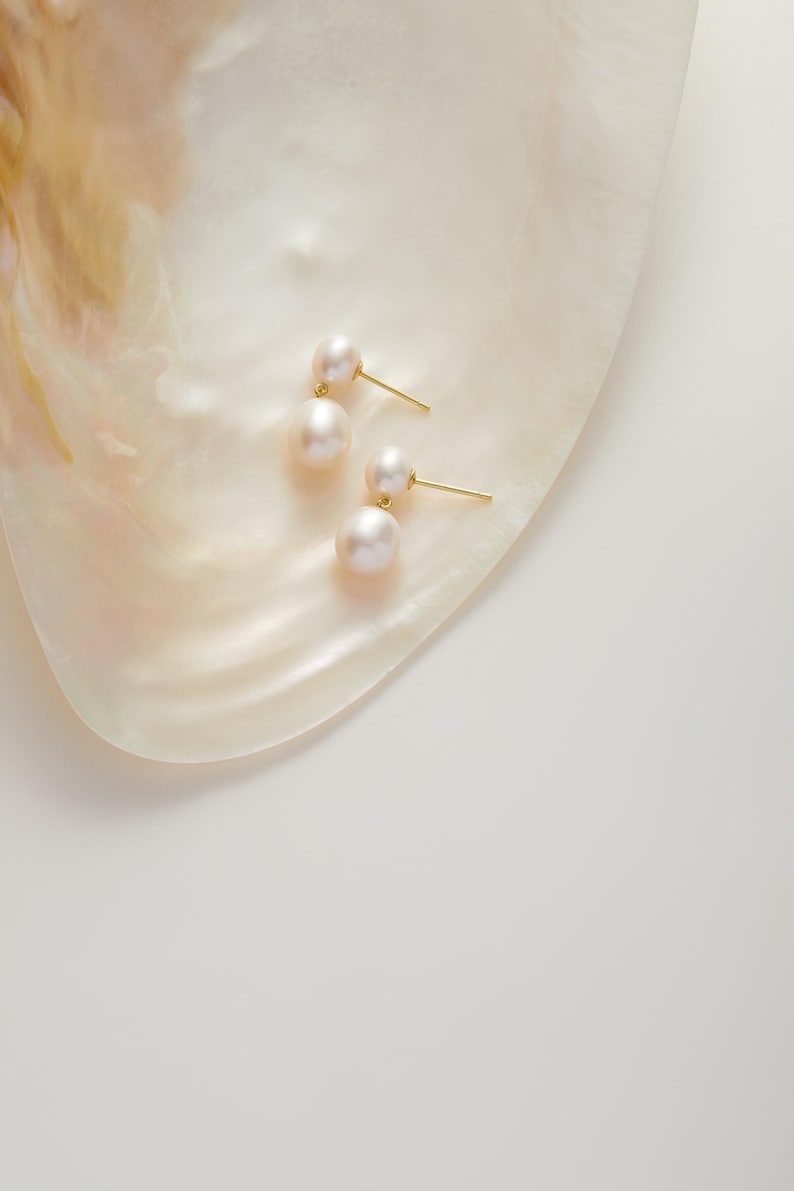 May include: Two pairs of pearl earrings with gold posts rest on a pearlescent, off-white surface. Each earring features two white pearls. The earrings are displayed on a shell-shaped surface, with a soft, diffused light.