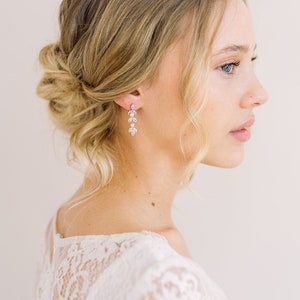 May include: Elegant dangle earrings with a floral design, featuring clear crystals. The earrings are worn by a person with blonde hair styled in an updo. The person is wearing a white lace top.