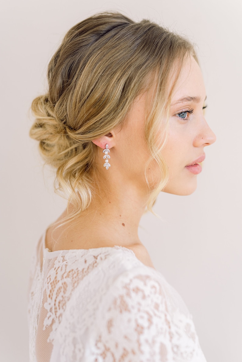 May include: Elegant dangle earrings with a floral design, featuring clear crystals. The earrings are worn by a person with blonde hair styled in an updo. The person is wearing a white lace top.